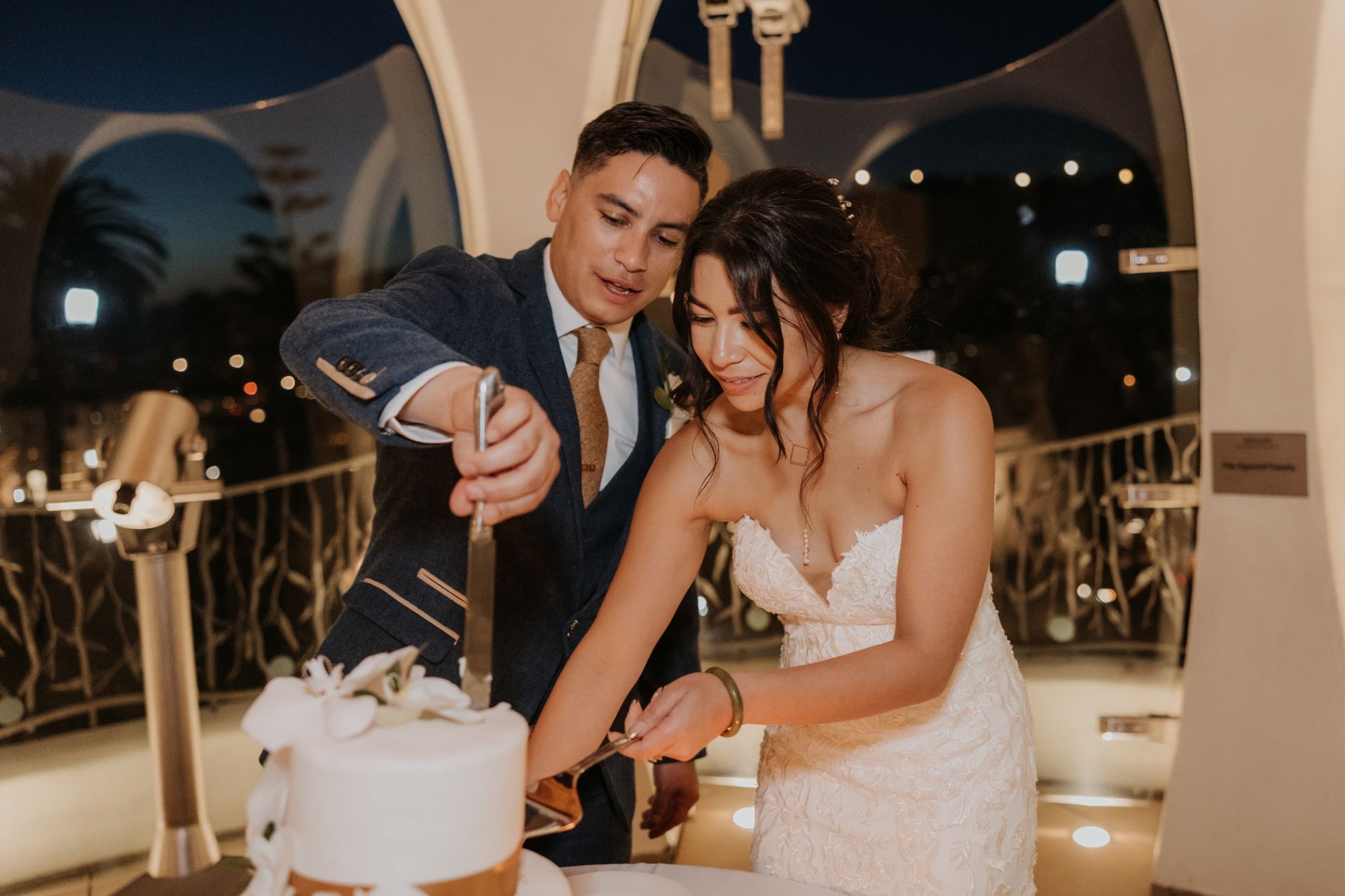 Couple cutting a white wedding cake indoors, smiling under warm lights.