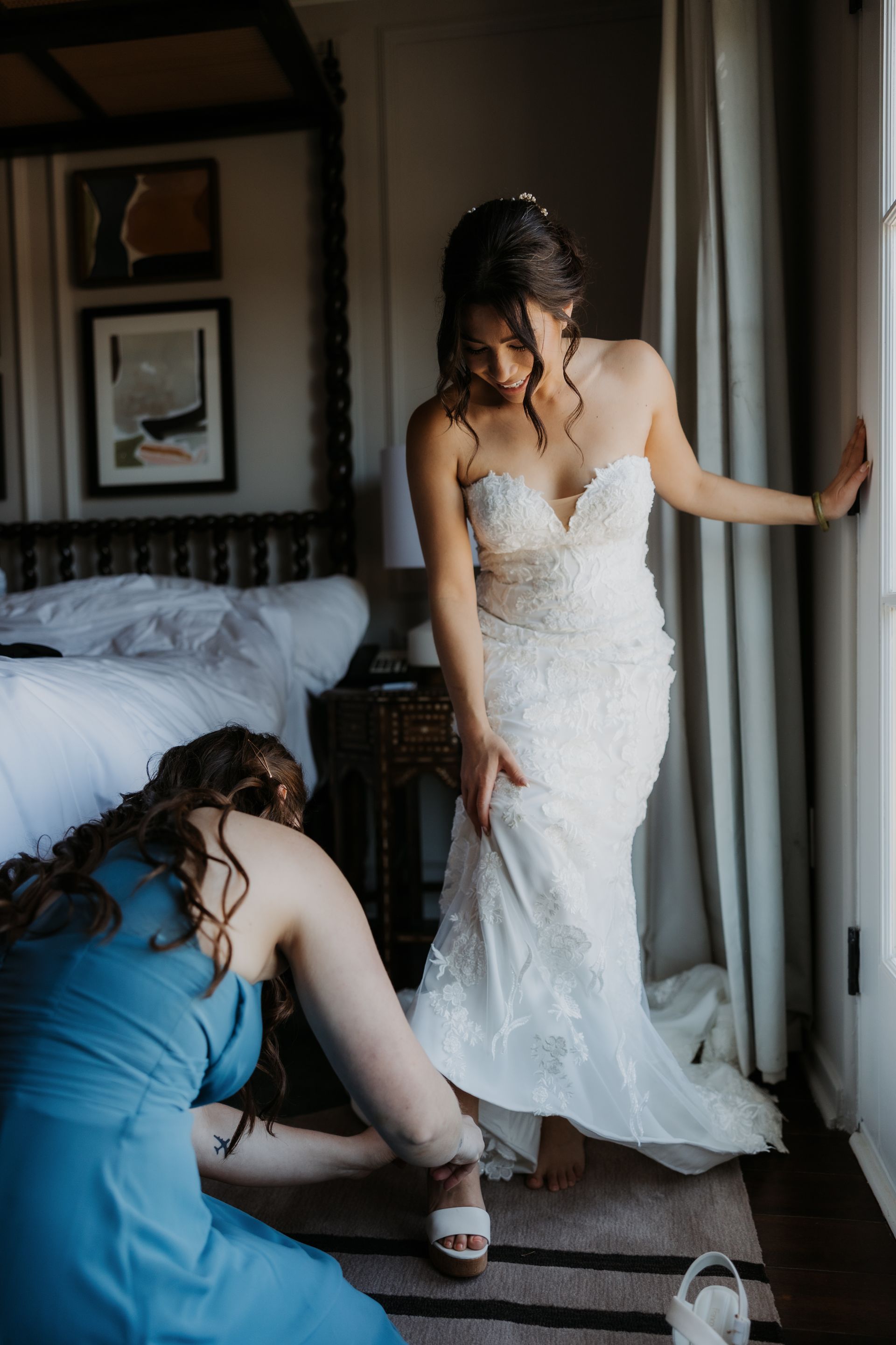 Bride in a white wedding dress standing by a window while another woman kneels to adjust her shoe