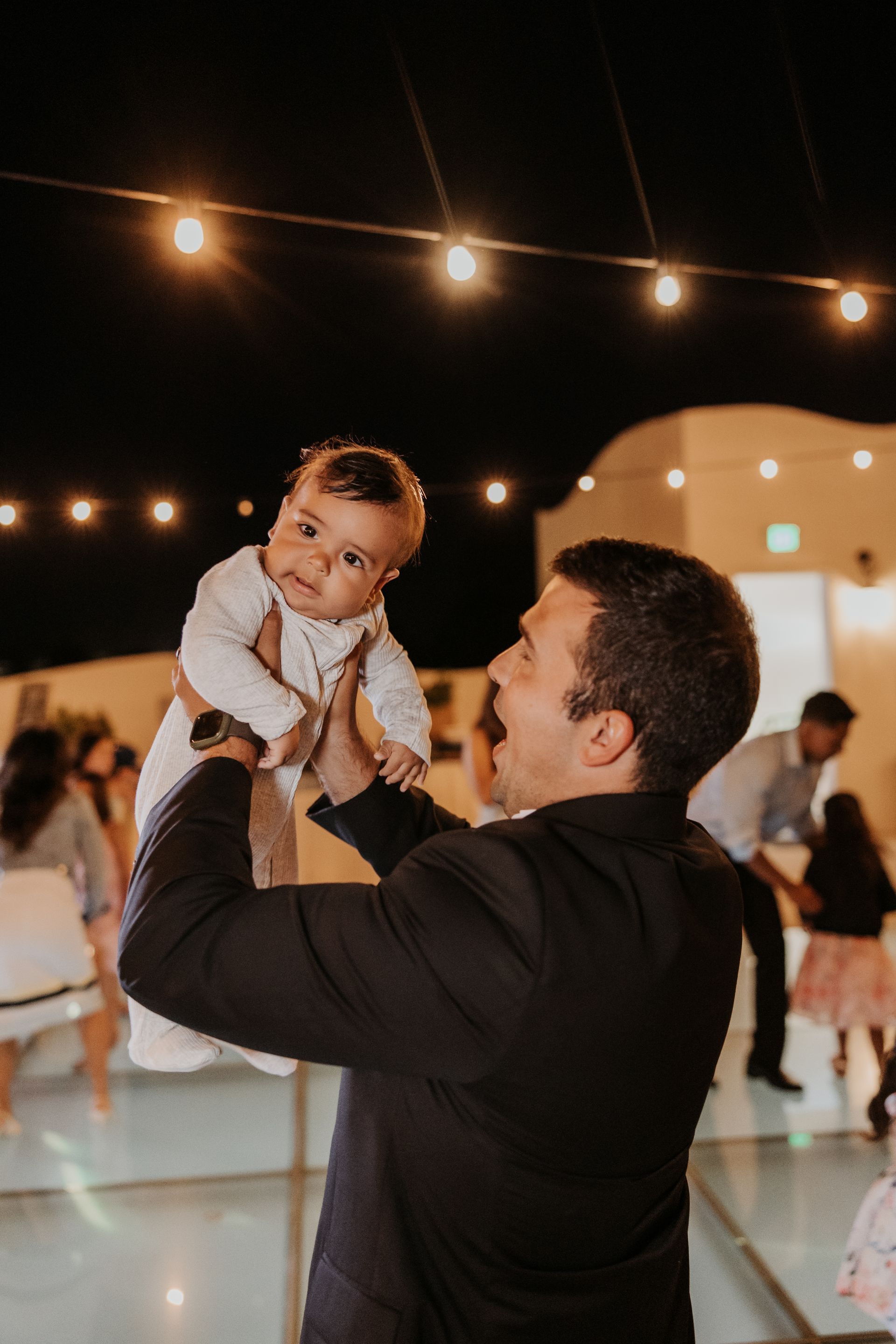 Man in a black suit lifts a baby at an outdoor evening party with string lights.