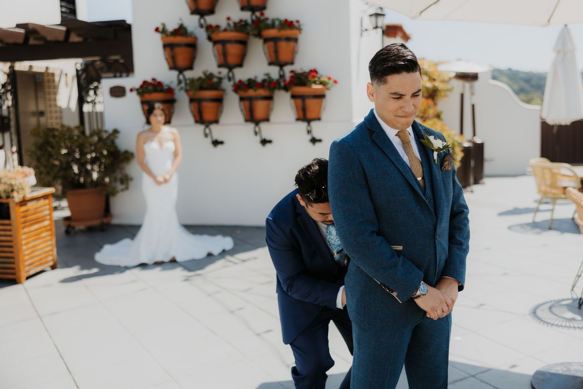 Man in blue suit and bow tie standing outdoors while another person adjusts his jacket at a wedding venue