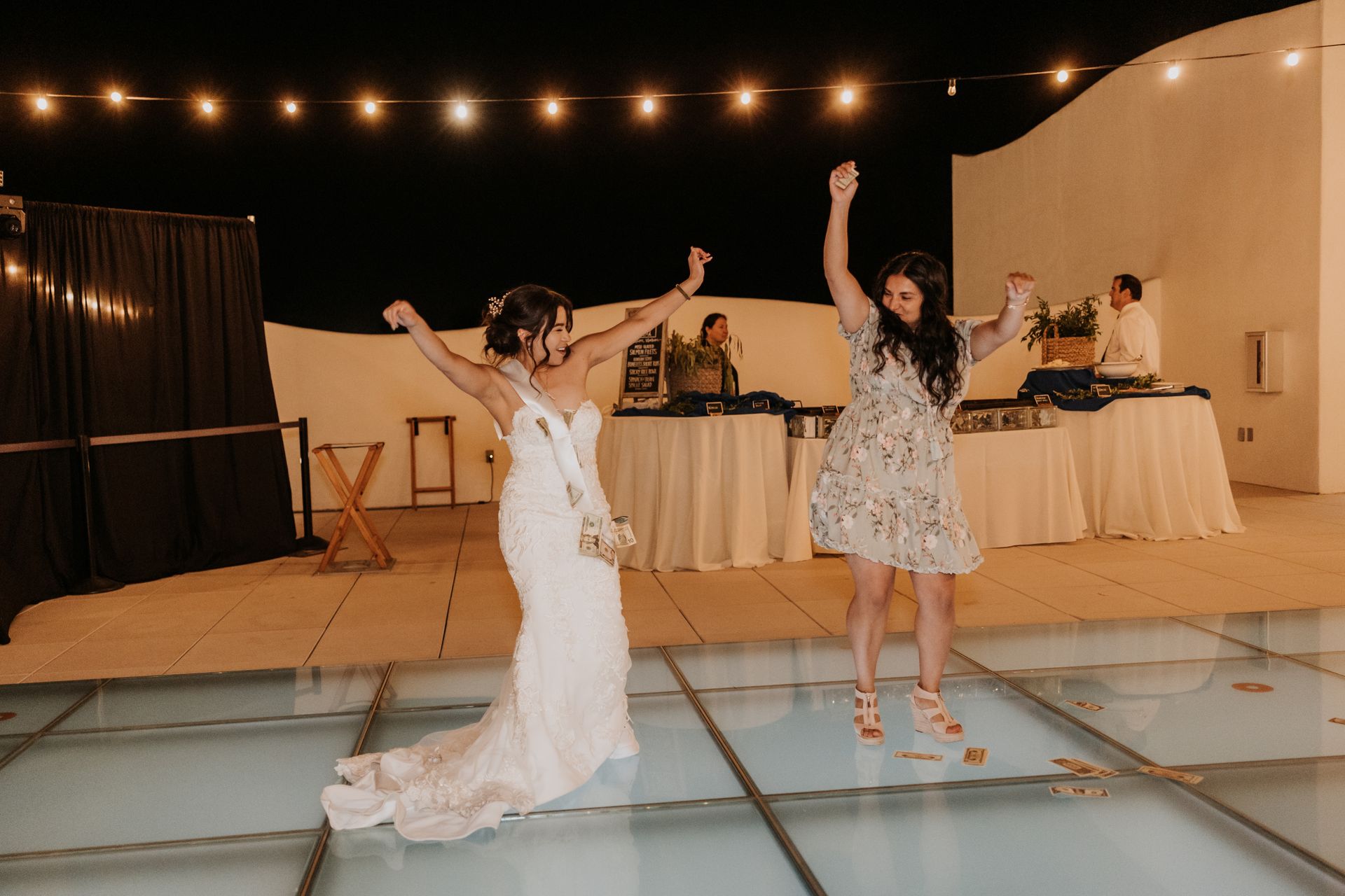 Two women dancing on a lit glass floor at an outdoor evening reception