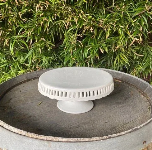 White round cake stand on a mesh-topped table outdoors, with green foliage behind it
