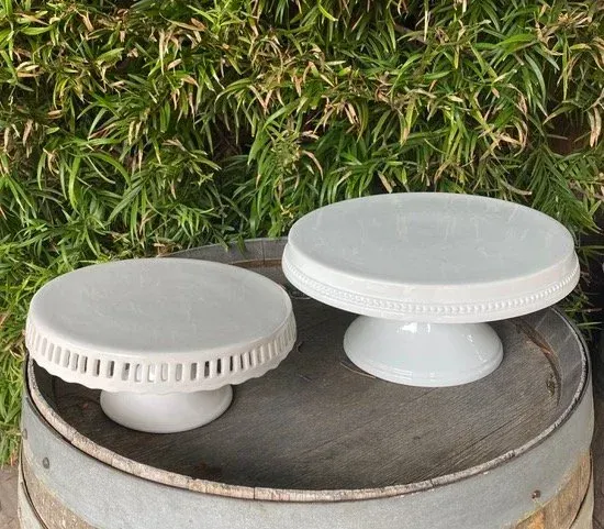 Two white round cake stands on a metal barrel in front of a leafy green wall