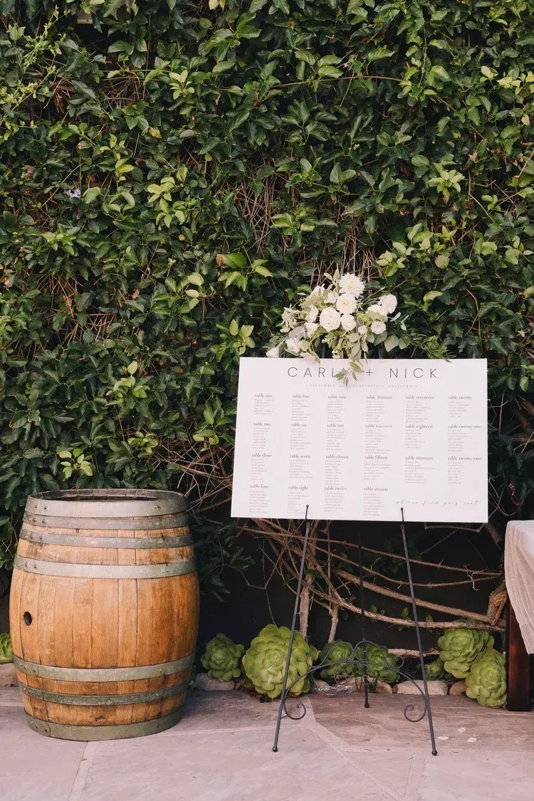 Outdoor event sign on a metal easel beside a wooden barrel, backed by a leafy hedge and flowers.