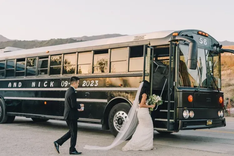 Bride and groom walking beside a black vintage bus on a road, wedding bouquet in hand