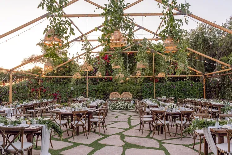 Outdoor reception venue with wooden tables, chairs, greenery, and a glass-roofed pavilion.