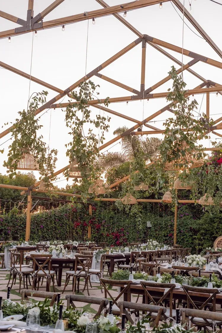 Outdoor café seating under a glass-roofed greenhouse with wooden beams and greenery.