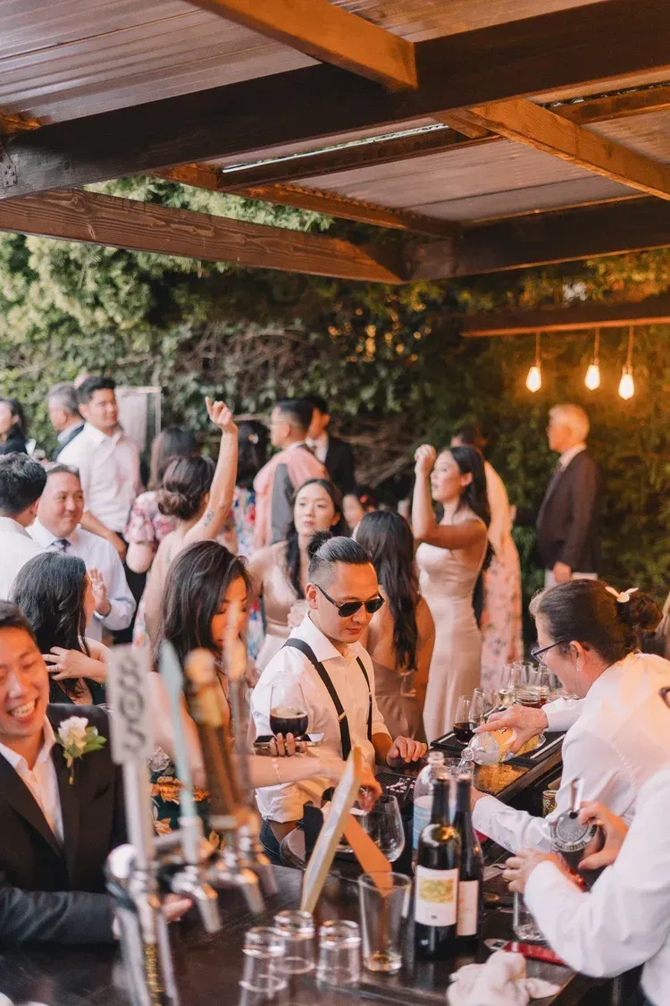 Guests mingle and toast at a lively outdoor evening reception under wooden beams and warm lights