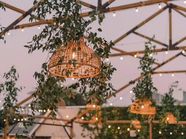 Warm string-lit patio with woven hanging lamps and leafy greenery under a geometric canopy