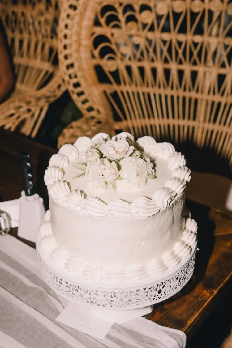 White frosted cake with flowers on a lace doily, set on a wooden table with wicker chairs behind it