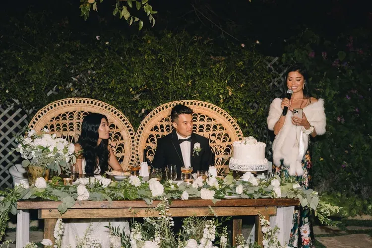 Outdoor wedding reception with bride and groom at a decorated table, seated beside a speaker