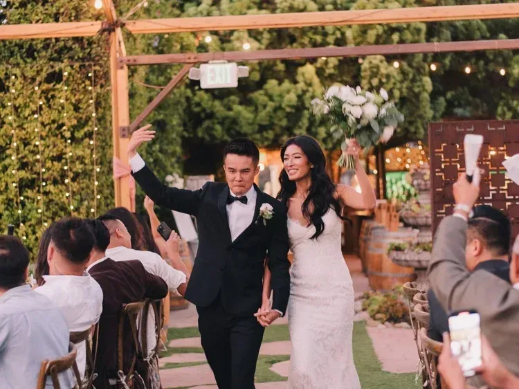 Newlyweds walk down an outdoor aisle, smiling and raising joined arms at their wedding ceremony.
