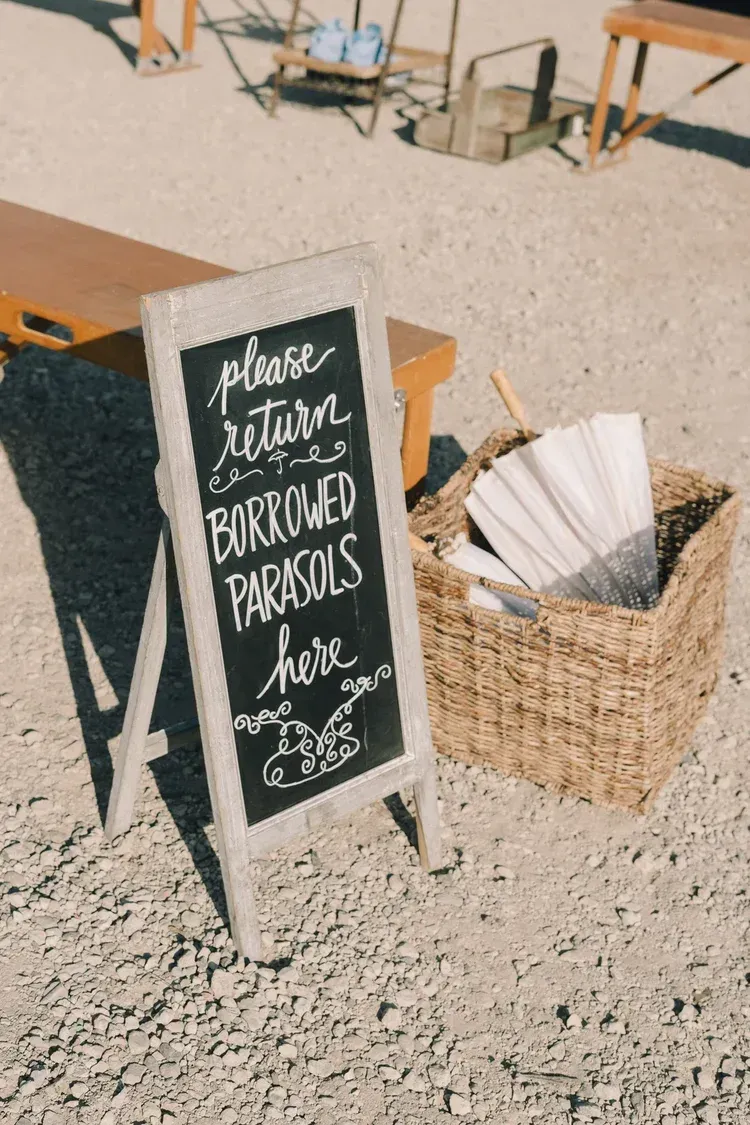 Wedding welcome sign in a woven basket on sandy ground, with parasols nearby