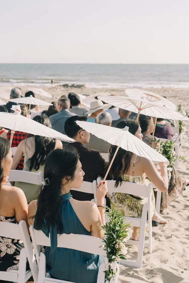 People seated on white chairs under umbrellas at a beach ceremony by the sea