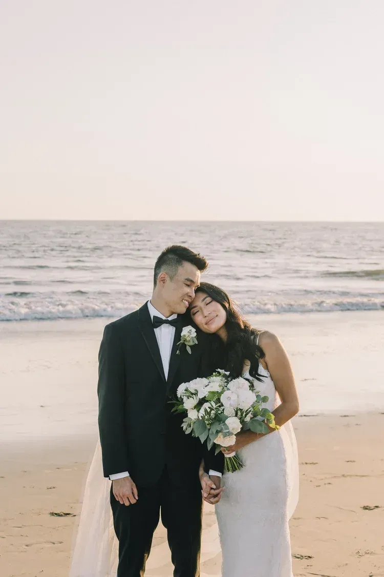Newlyweds in black tuxedo and white dress embracing on a beach at sunset, holding a bouquet