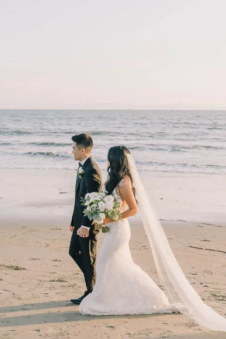 Bride and groom standing on a beach with waves in the background, bride holding a bouquet and veil trailing