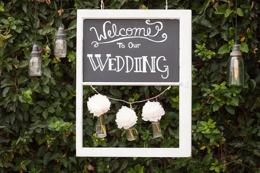 Wedding welcome sign with white frame and hanging flowers against a green leafy background