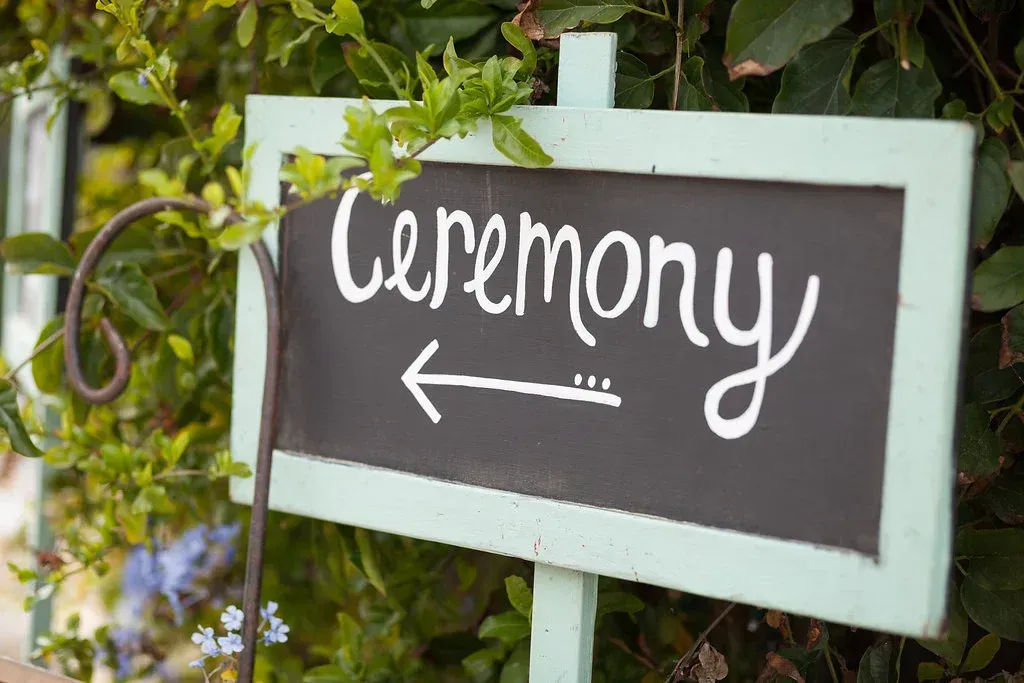 Sign pointing left to a ceremony, with white handwritten text on a dark board