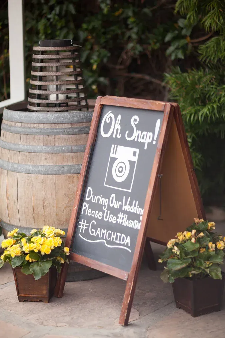 Small café sidewalk sign beside a wooden barrel and yellow flowers, with a handwritten menu board.