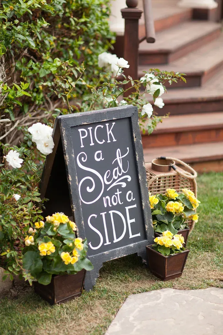 Chalkboard sign reading “Pick a seat not a side” beside yellow flowers and white blooms outdoors