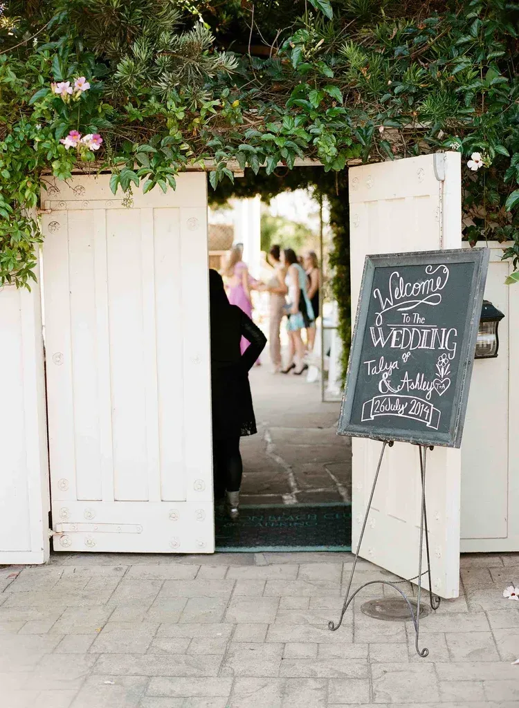 Open white gates with a chalkboard sign at a garden entrance, with people visible inside.