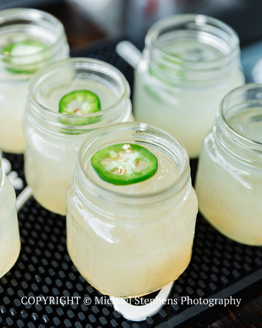 Mason jars of pale drink topped with jalapeño slices on a black tray