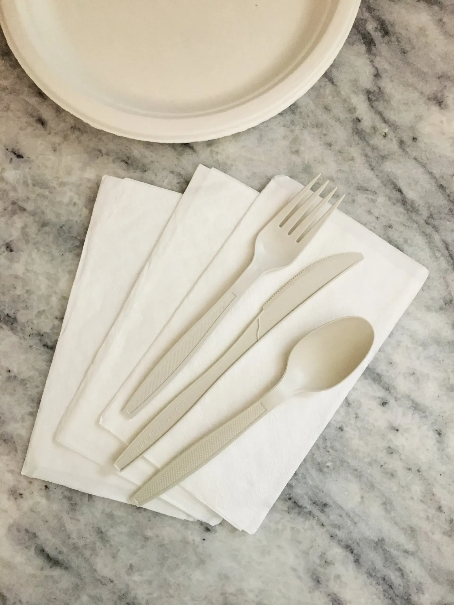 Paper napkins with a fork and spoon on a marble tabletop, beside a plate.