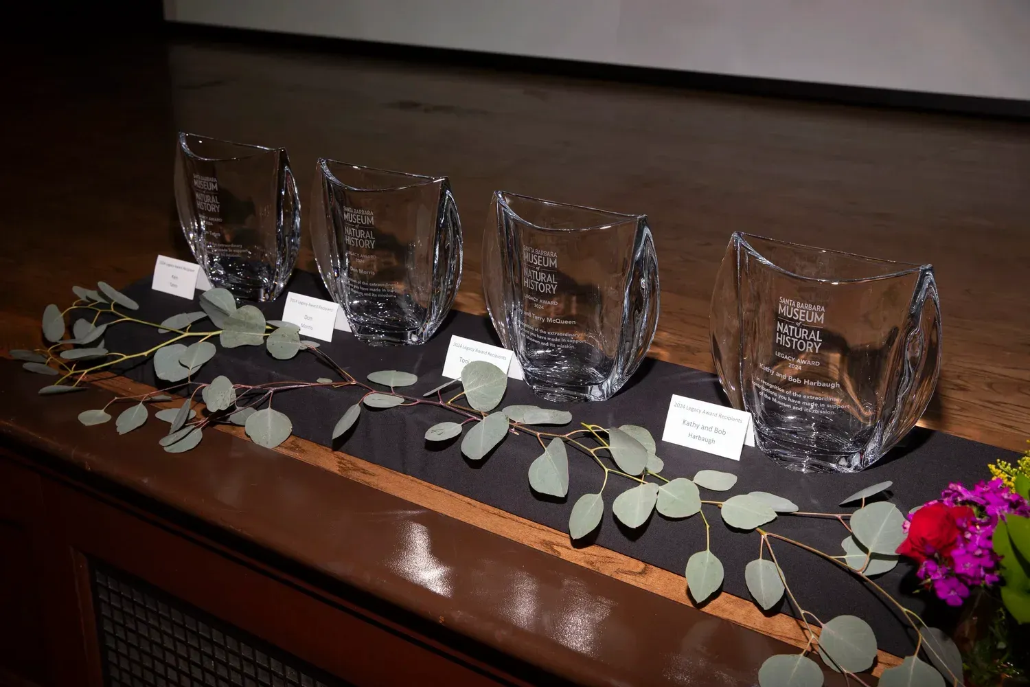 Five crystal award vases with white tags on a polished wooden table, decorated with leaves and flowers.