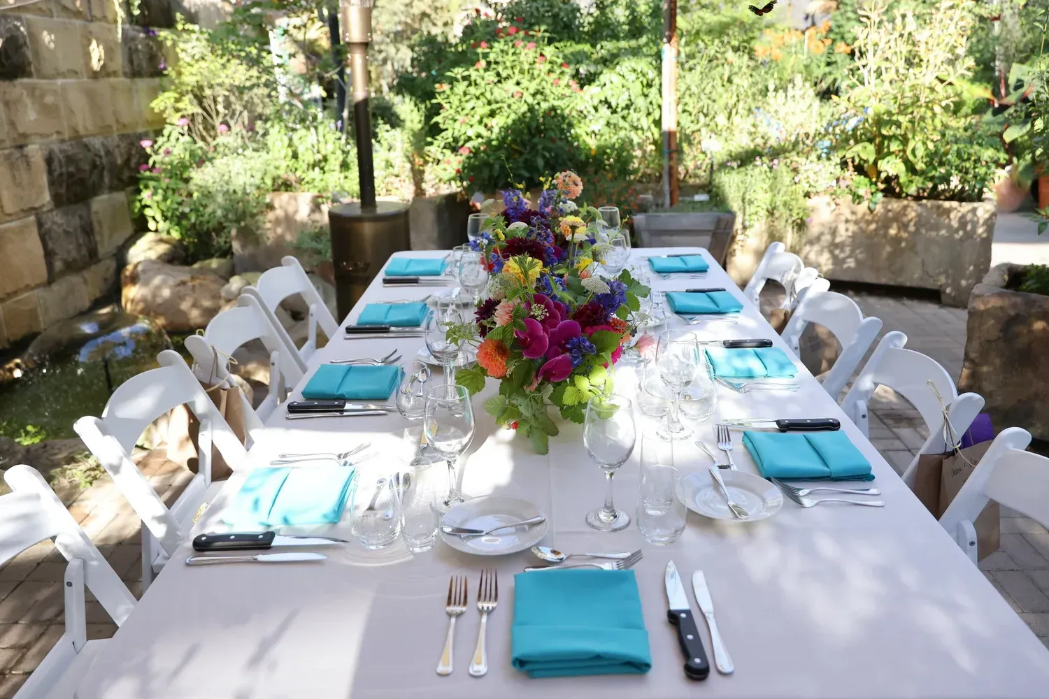 Long outdoor dining table with turquoise napkins and a colorful floral centerpiece in a garden patio