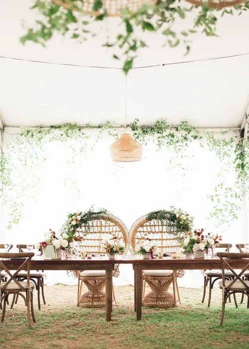 Elegant outdoor dining setup under greenery with rustic wooden tables and chairs on grass.