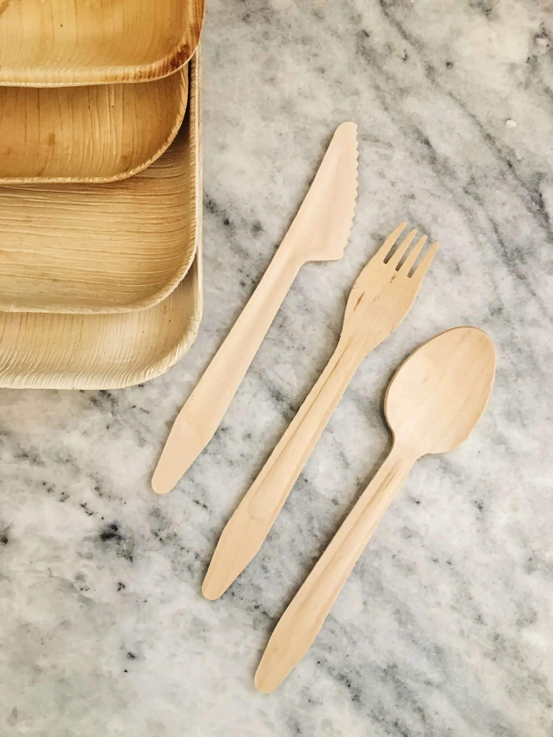Wooden reusable knife, fork, and spoon beside stacked bamboo plates on a marble surface