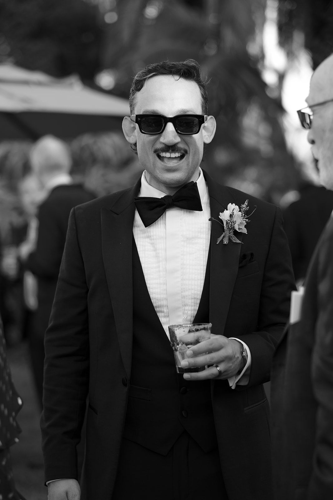 Man in a tuxedo and sunglasses holding a drink at an outdoor event
