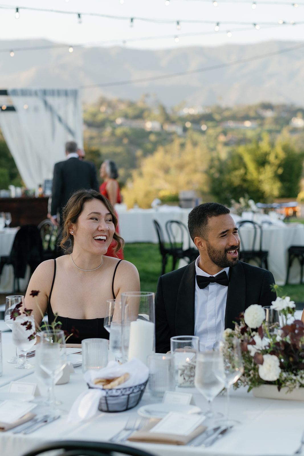 Smiling couple seated at a decorated outdoor wedding reception table with mountains in the background