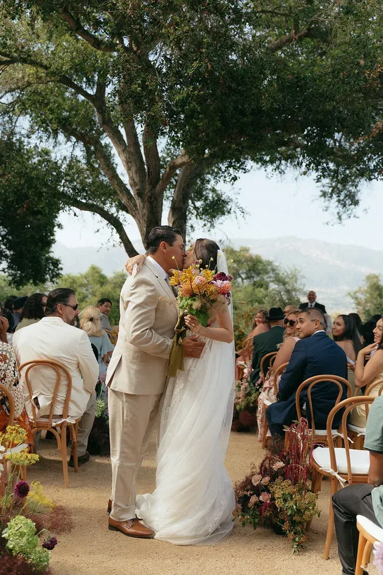 Bride and groom kiss at outdoor wedding under a large tree, guests seated nearby.