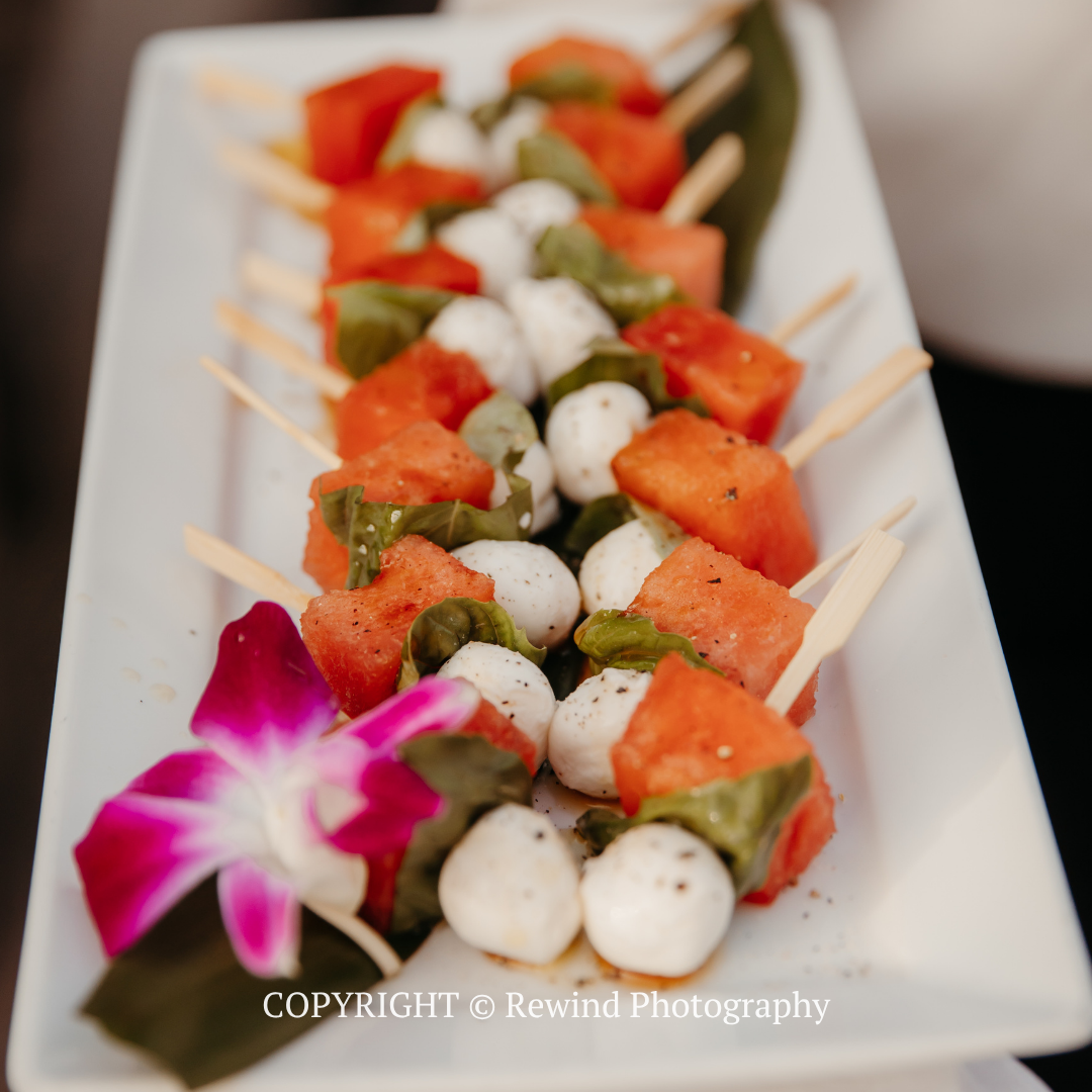 Skewers of watermelon, cucumber, and mozzarella on a white platter, garnished with a purple flower.