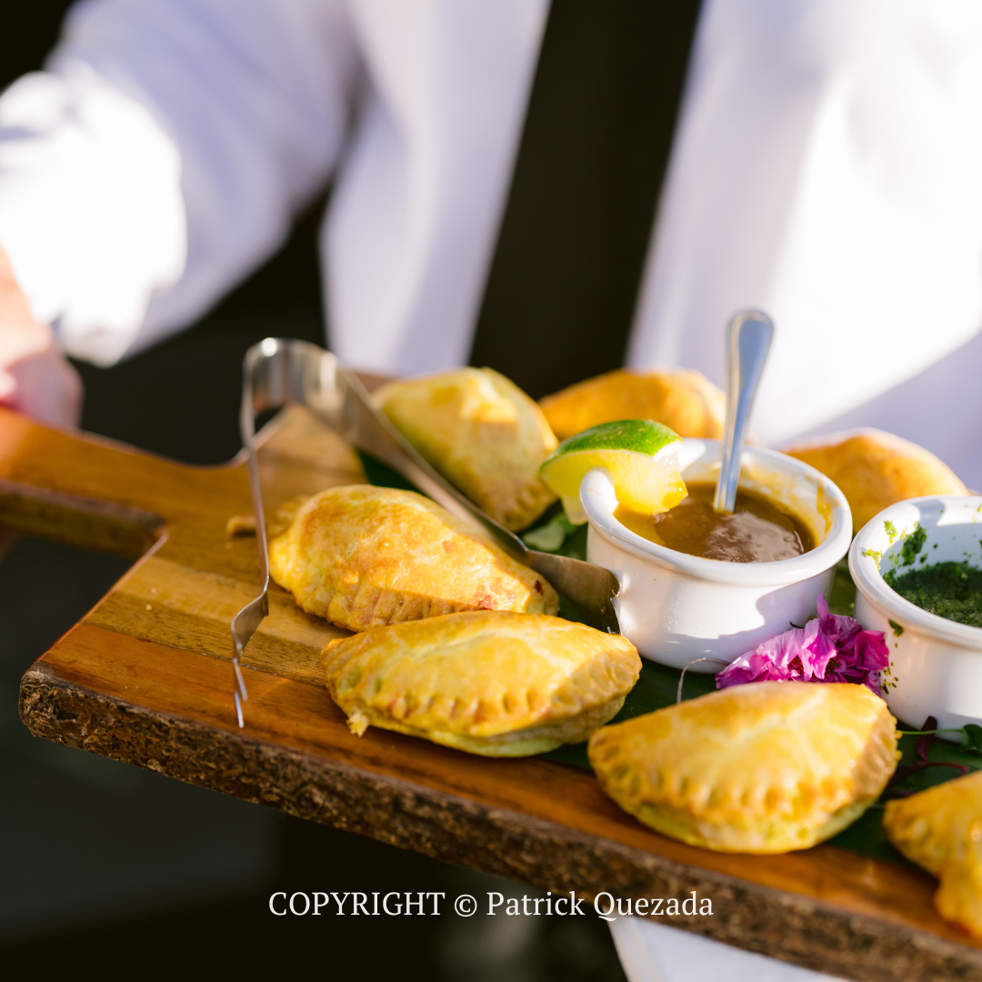 Assorted empanadas on a wooden board with dipping sauce and garnish, served by a person in a white jacket