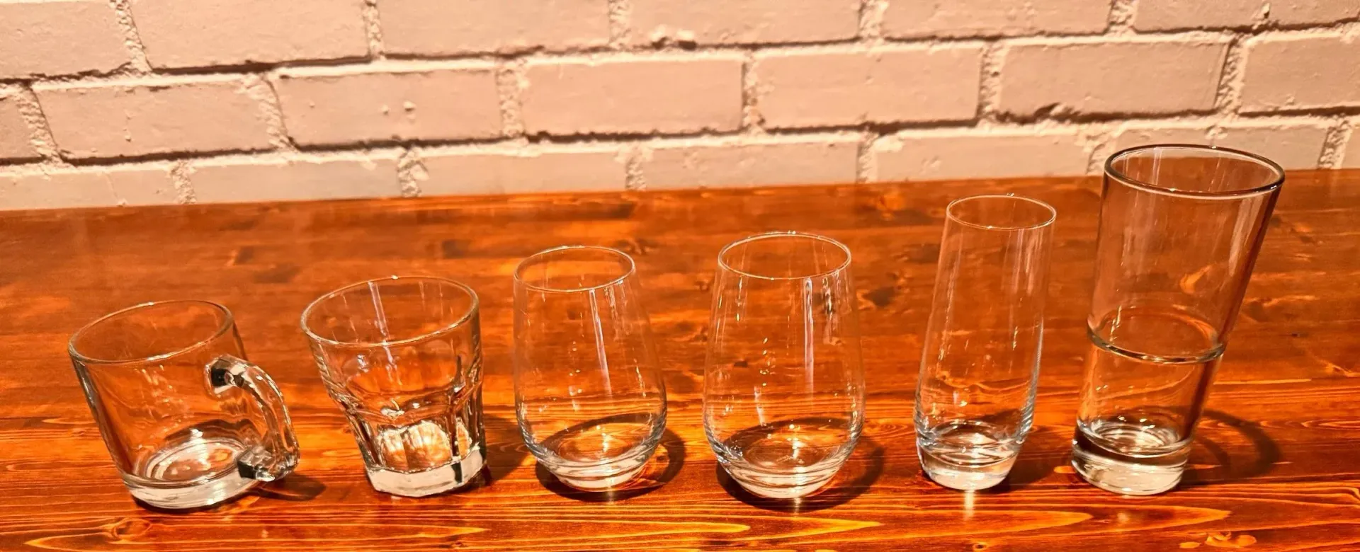 Row of six clear drinking glasses on a wooden table against a brick wall