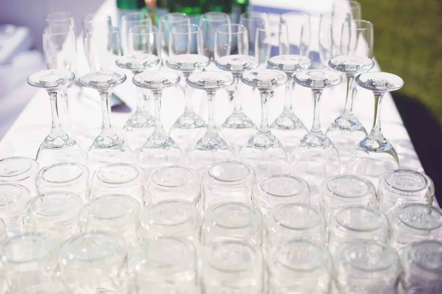 Rows of clear wine glasses and tumblers arranged on a white tablecloth at an event setup