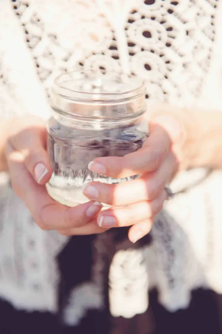 Hands holding a small glass jar of water in front of a white crocheted top.
