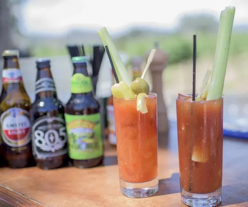 Two Bloody Mary cocktails with celery and olives beside beer bottles on a wooden table outdoors