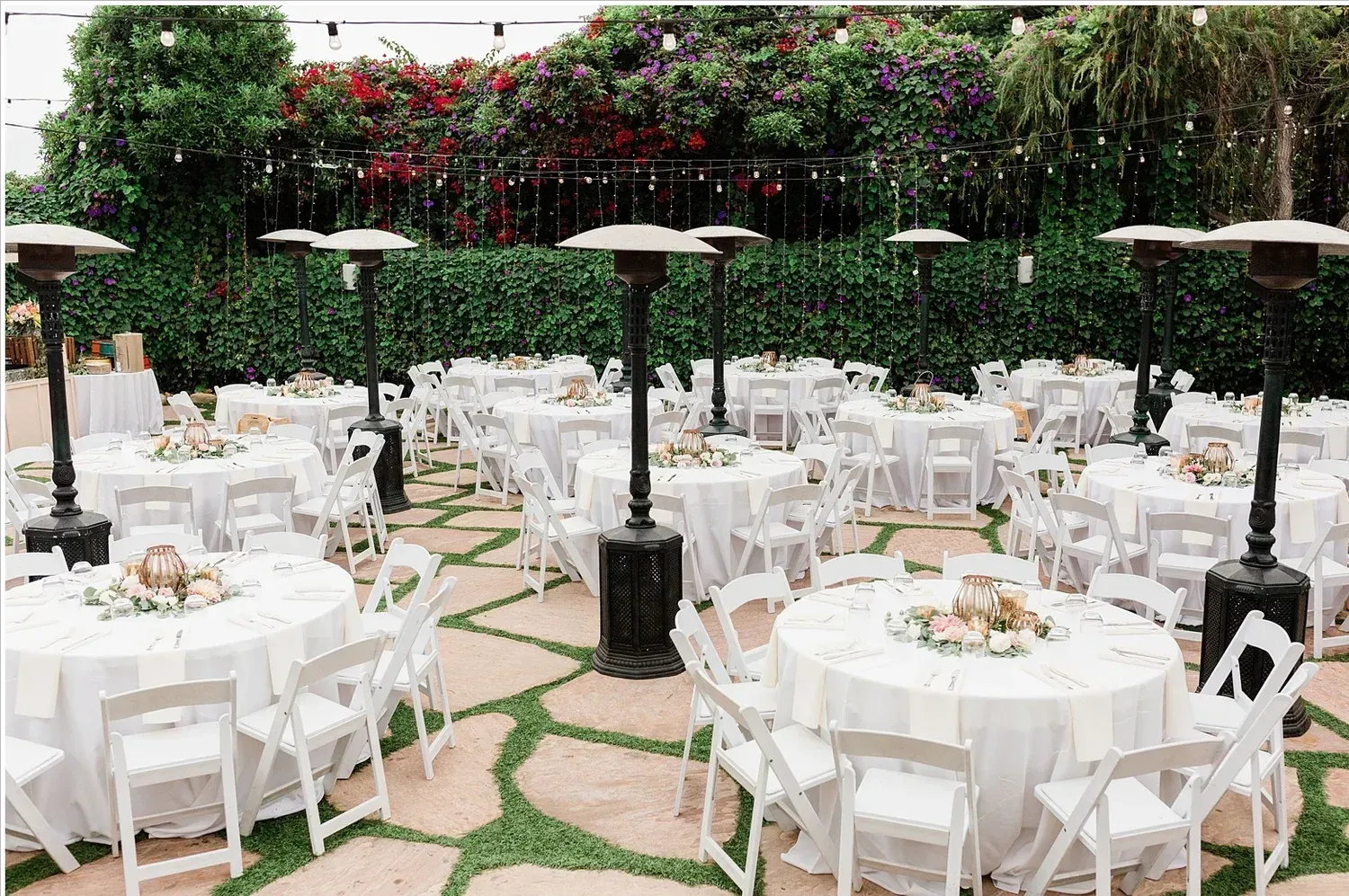 Outdoor reception setup with white round tables, chairs, and tall heaters beneath green hedges and string lights