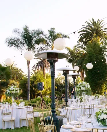 Outdoor event setup with white chairs, round tables, and tall patio heaters among palm trees at dusk