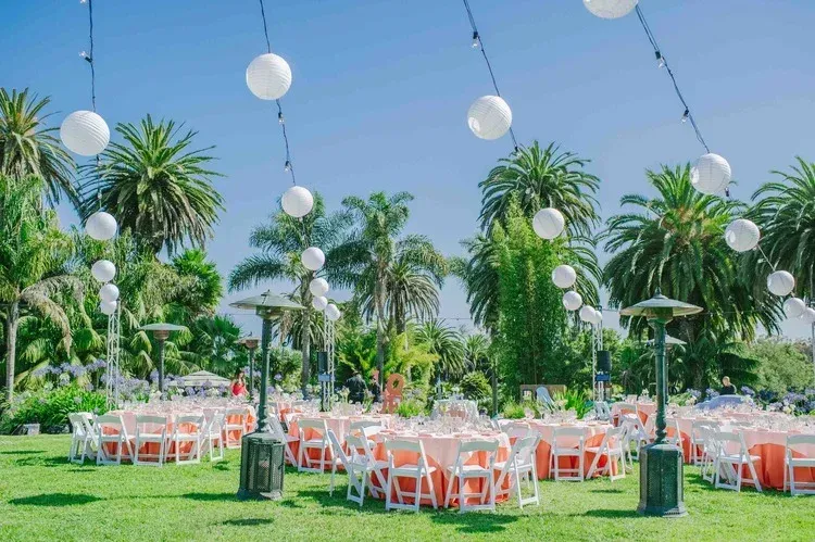 Outdoor event setup with white chairs and tables under hanging lanterns in a lush garden.