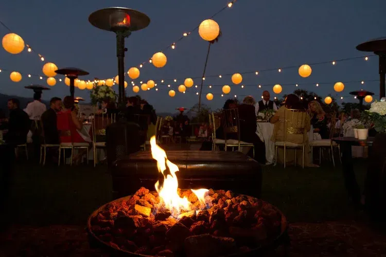 Outdoor evening gathering with lanterns and a fire pit, people seated at tables in the background