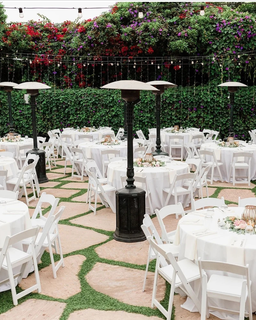 Outdoor event patio with white tables and chairs, black heaters, and lush floral greenery overhead.