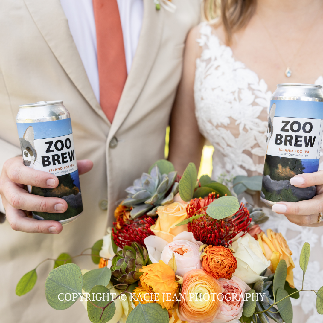 Two people in formalwear hold ZOO BREW cans beside a colorful fruit-topped cake.
