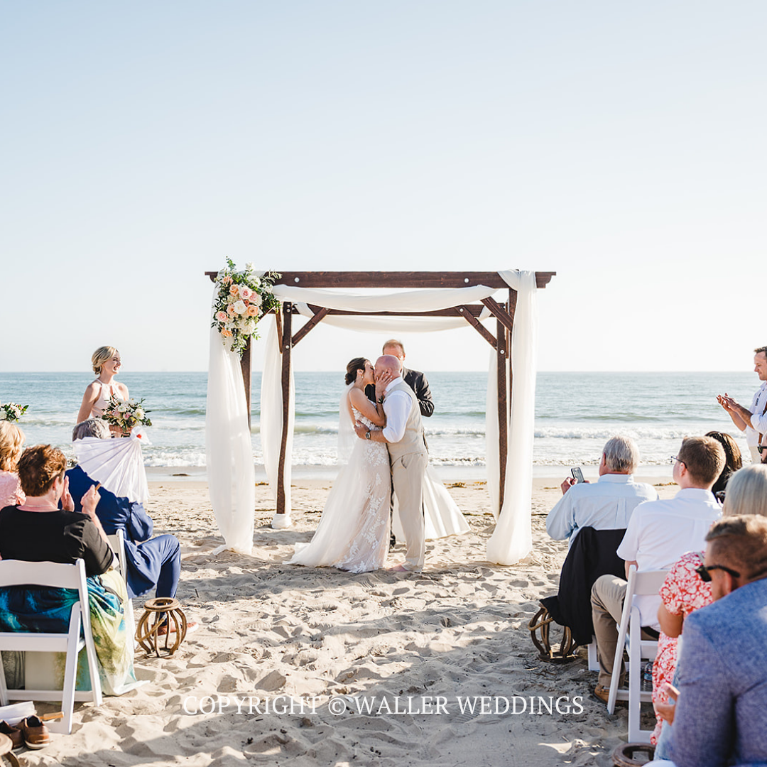 Beach wedding ceremony under a wooden arch with white drapes, bride and groom kissing, seated guests nearby