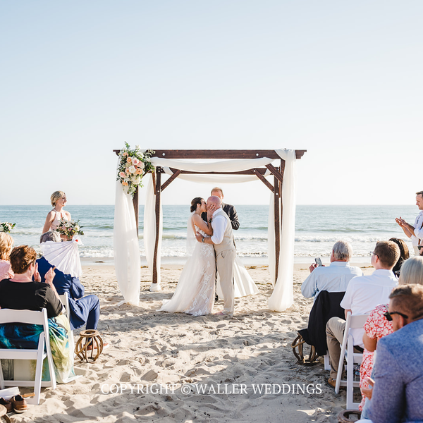 Bride and groom kissing under a beach wedding arch as guests watch from white chairs.