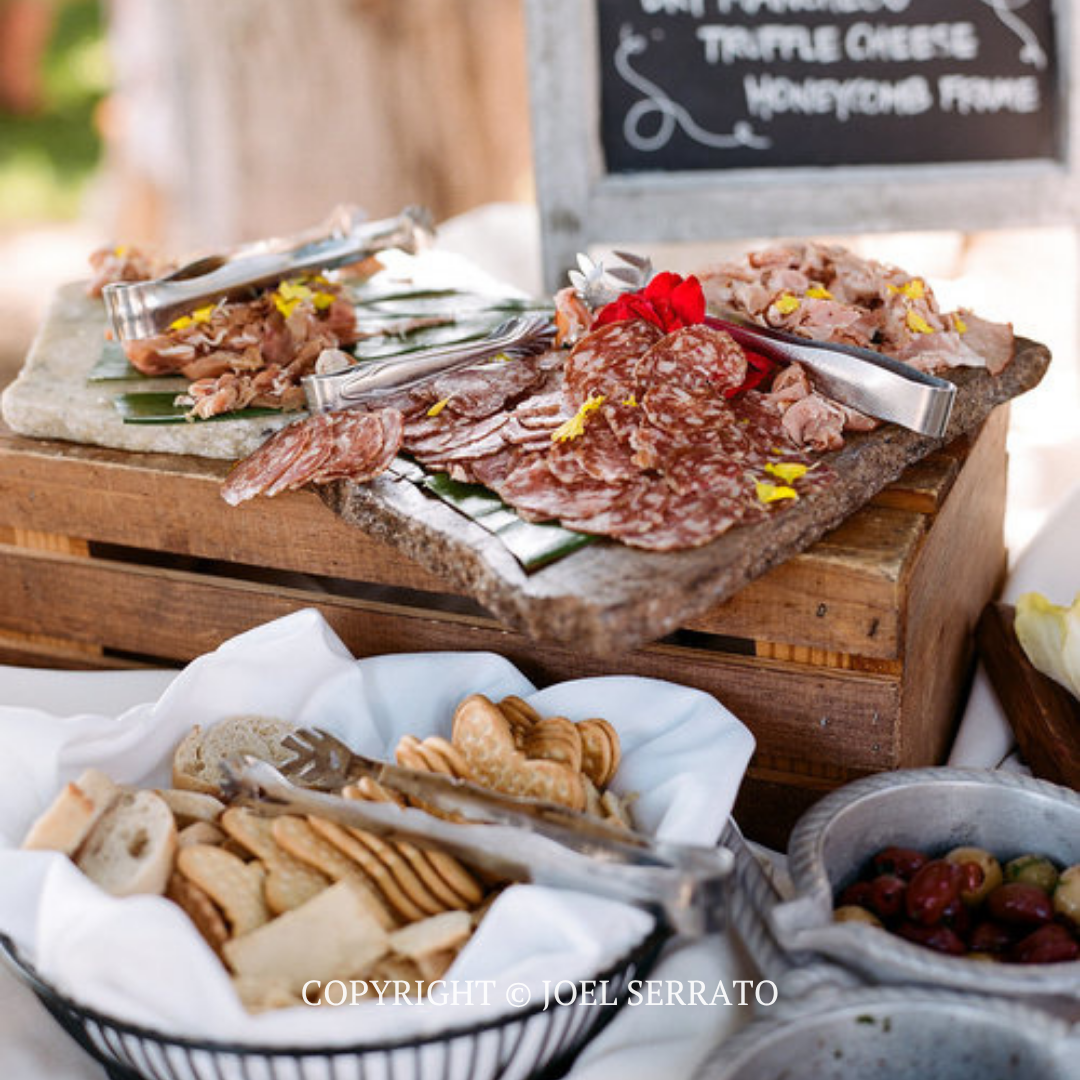 Assorted cured meats and cheese on wooden boards at an outdoor buffet, with bread and crackers below