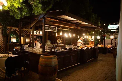 Nighttime outdoor bar with warm string lights, wooden counter, and barrels in a courtyard